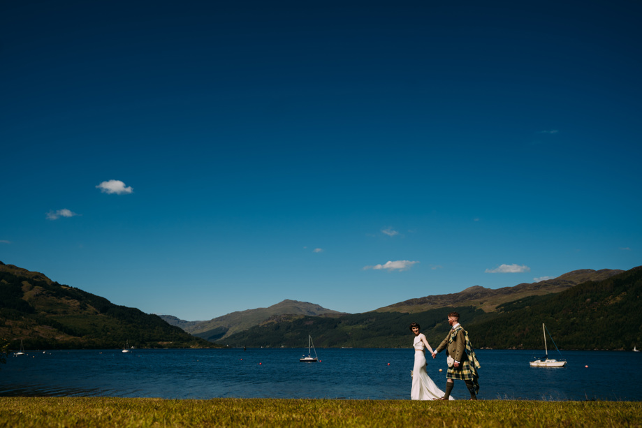 Bride and groom hold hands whilst walking along waterside with boats in background